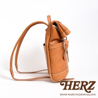 職人手作淺棕色皮革雙口袋折蓋後背包，採用真皮材質，側邊可見一處口袋，包身有縫線細節，配有可調整式背帶。