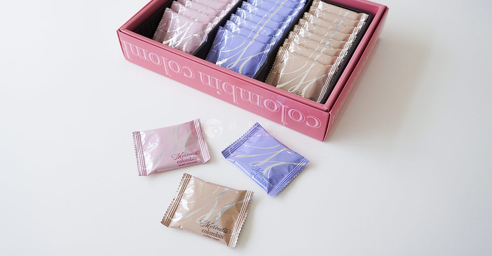 粉紅色禮盒裝著 Merveille Colombin 綜合巧克力夾心餅乾，有粉色、紫色和米色三種包裝，其中三塊單獨的餅乾放在禮盒旁，分別是粉色、紫色和米色包裝。