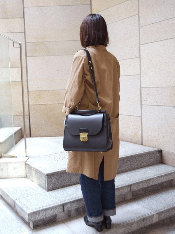 一名女子身穿卡其色外套，側背黑色皮革製的兩用單肩包，包款帶有屋瓦造型翻蓋與金色鎖扣。