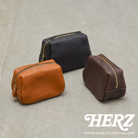 三個日本HERZ手工製作的Organ系列皮革收納包置於淺灰色平面上，分別呈現焦糖色、黑色與深咖啡色，包身外型簡約圓潤並配有金色拉鍊，右下角印有HERZ品牌標誌與手工製造年份字樣。