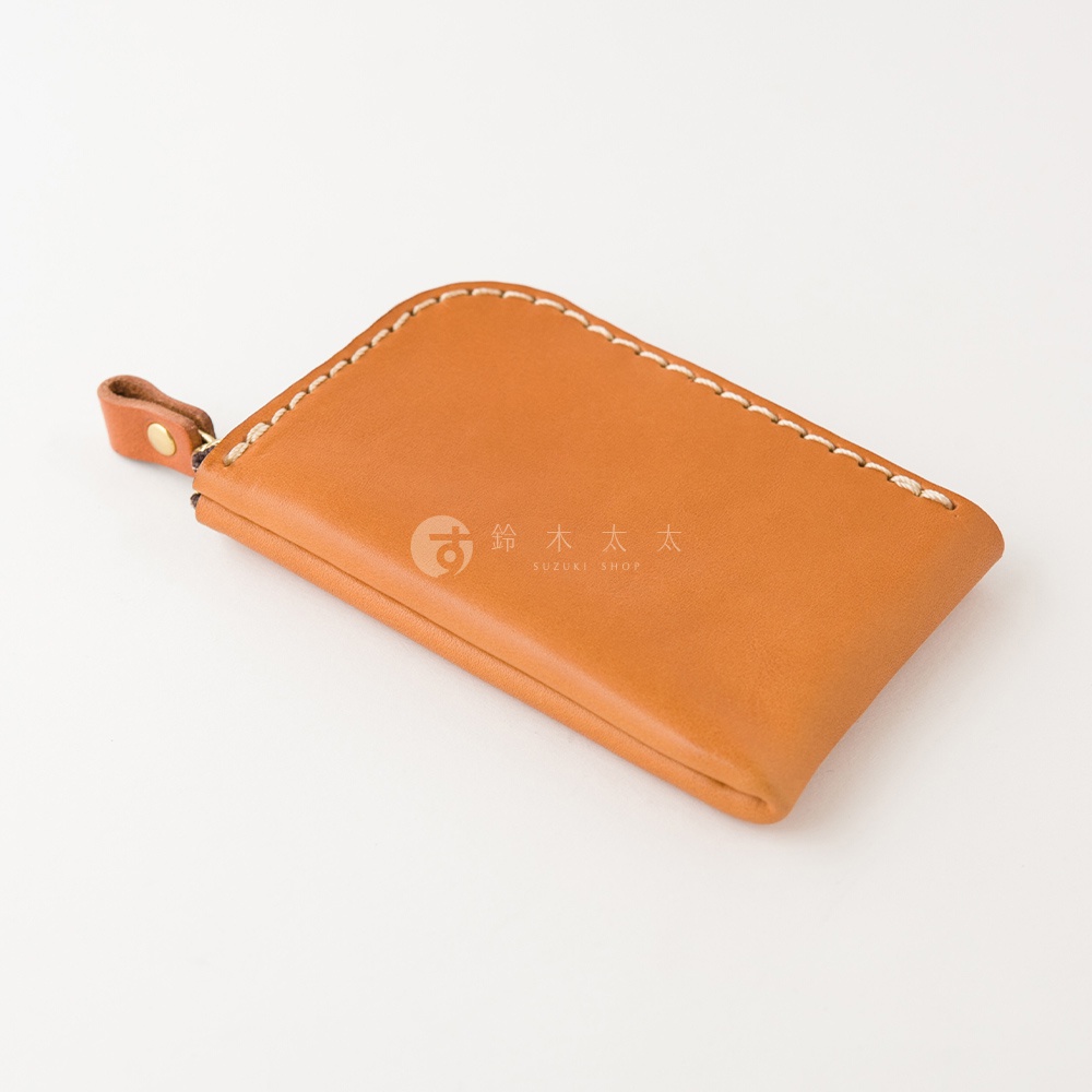 一個擺放在純白背景上的日本HERZ職人手作皮革零錢包，材質為Stare皮革，呈現溫潤的棕色調，整體設計簡約精緻，側面配有皮革拉鍊環與金色金屬扣細節，並以手工縫線固定邊緣，視覺上呈現出優雅且紮實的皮革質感。