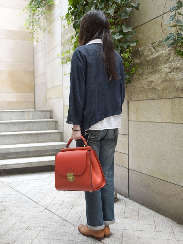 一款橘紅色皮革手提包，具有瓦片狀的立體設計，搭配金色鎖扣和提把，可作為單肩包使用。