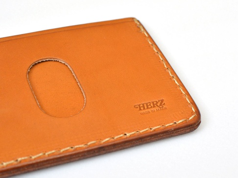 職人手作皮革橫式票券收納夾，採用天然皮革製成，顏色為溫暖的棕褐色。表面有壓印的「HERZ MADE IN JAPAN」字樣，以及一個用於放置票券的橢圓形開口。縫線整齊，邊緣處理細緻。
