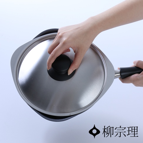 一隻手拿著蓋著不鏽鋼蓋的柳宗理岩紋感單手鐵鍋，鐵鍋呈特殊不對稱形狀，鍋身為深灰色鐵質表面，搭配黑色手柄與黑色圓形鍋蓋頭，背景為素雅的純白色。