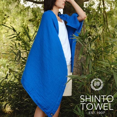 一位女性身披一件寶藍色2.5層紗質地暖毯，暖毯柔軟且帶有細緻織紋，背景為綠意盎然的樹林。