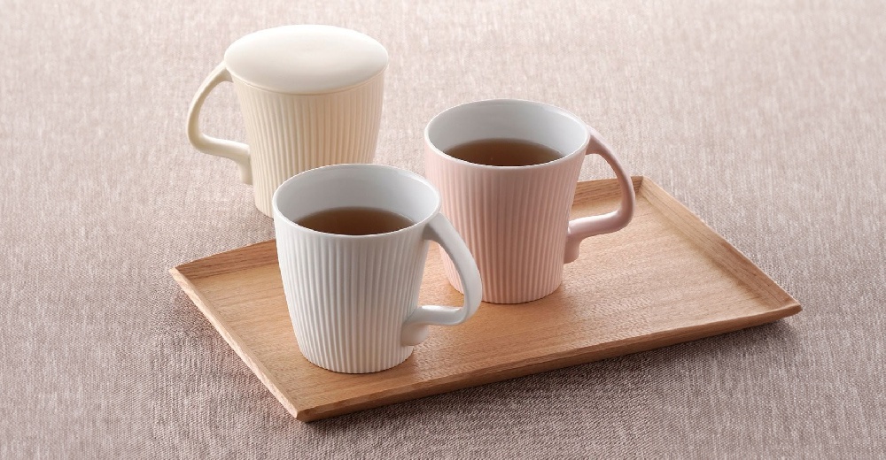 木質托盤上擺放著三個馬克杯，其中兩個杯子內有茶水。杯子為陶瓷材質，外觀有垂直的細長紋路，提供白色、粉紅色和米黃色。其中一個米黃色杯子配有同色系杯蓋。