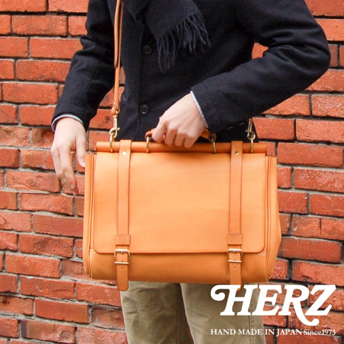日本HERZ職人手作皮革屋瓦風側背包，以橘棕色皮革製成，配有金色五金件和可調節的肩帶。