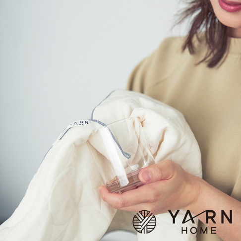 手持一杯水，將其靠近一件米白色、帶有菱形格紋縫線的廚房拭巾。拭巾邊緣有灰色滾邊，並縫有「YARN HOME」品牌標籤。