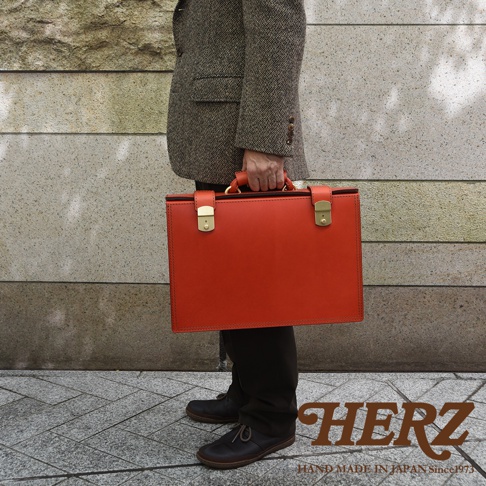 亮橘色職人手作皮革機師箱，配有黃銅色鎖扣及提把，展現經典設計。