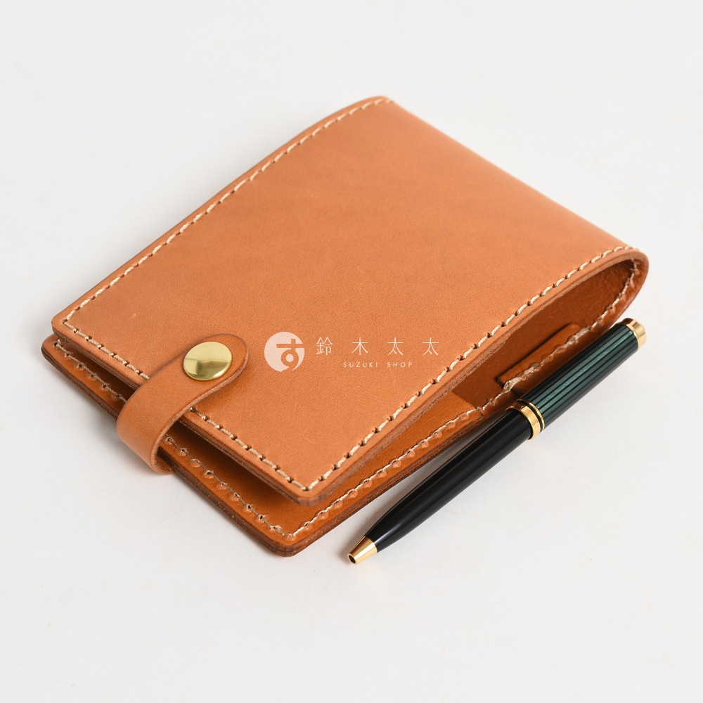 職人手作皮革便利貼收納套，淺棕色皮革，搭配黃銅鈕扣，縫線清晰，一旁放置一支綠色條紋金屬筆。