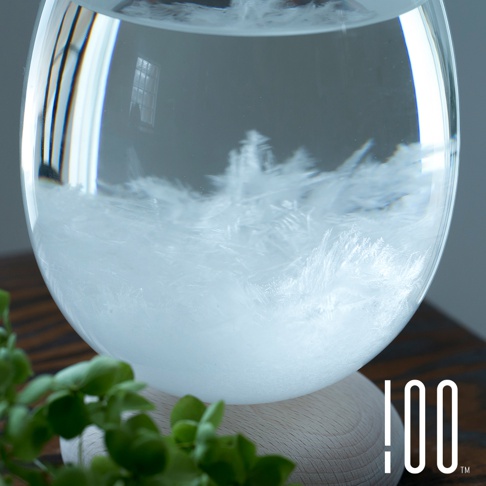 一個透明玻璃製成的圓形天氣瓶，內含純淨的水，瓶底沉澱著如同雪花般細緻的白色結晶。瓶子放置於一個淺木色的圓形木製底座上。