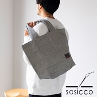 一款採用三河木綿刺子織材質的黑色與白色格紋大容量托特包，由一位男性從側面背起，包包上有一個紅色小標籤。