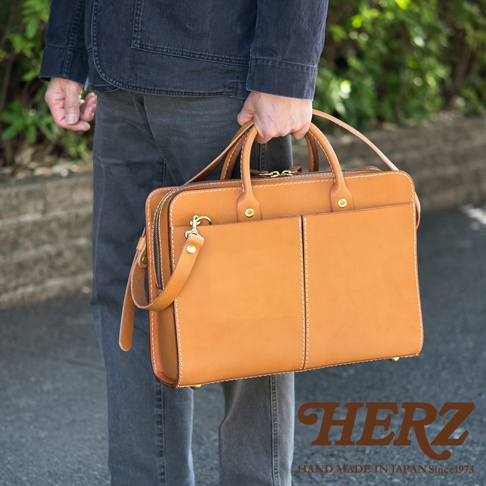 一位男士提著一個棕褐色皮革雙口袋兩用商務公事包，包包有兩個主要隔層，正面有兩個方形口袋，並配有頂部提把和可拆卸肩帶。