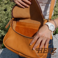 HERZ 品牌皮革大容量馬鞍包，近看展示包包內部夾層與精緻縫線，一位女性的手戴著白色手錶和銀色手鍊，正在整理包包，背景是綠色植物。