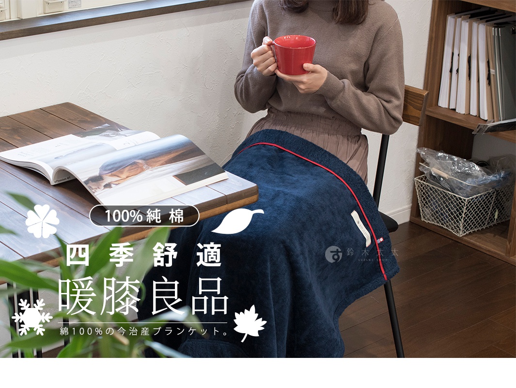 深藍色觸感柔軟的膝蓋毯，邊緣飾有紅色滾邊，上面有白色葉子圖案及品牌標籤。毯子覆蓋在一位女士的腿上，她正坐在椅子上，手持紅色杯子。