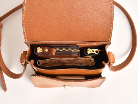 職人手作皮革翻蓋迷你單肩包 (CP-6)，採用柔軟的棕色皮革製成，設有兩個拉鍊隔層，內含一個小錢包、一本筆記本和一支手機。包款配有可調節的皮革肩帶，整體風格簡潔實用。