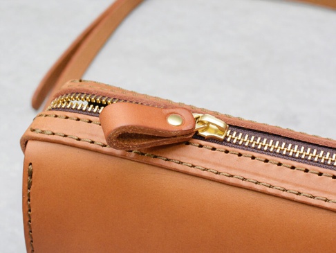 職人手作皮革單肩包，採用天然棕色皮革，配有金色金屬拉鍊和皮革拉頭，車縫線清晰，風格簡潔。