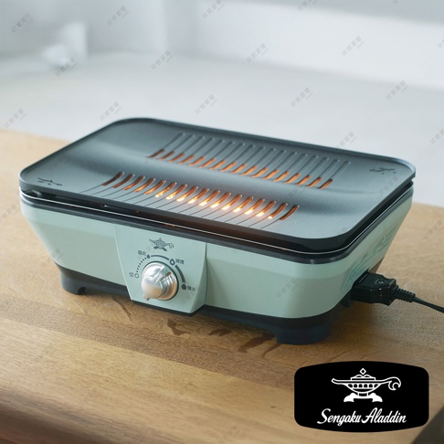 淺綠色迷你烤盤，配有黑色烤網，網格下方有橘色火焰，側面有銀色旋鈕和白色標示，底部有黑色防滑墊。