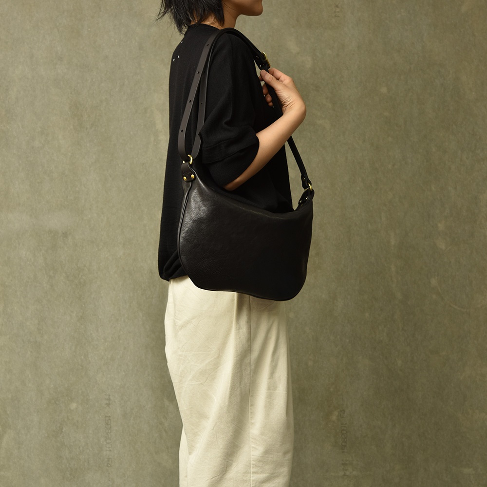 一位模特兒身著黑色上衣和米白色長褲，側身展示一個黑色皮革單肩半月包。包包採用柔軟的皮革材質，配有可調節的肩帶和金屬扣環。