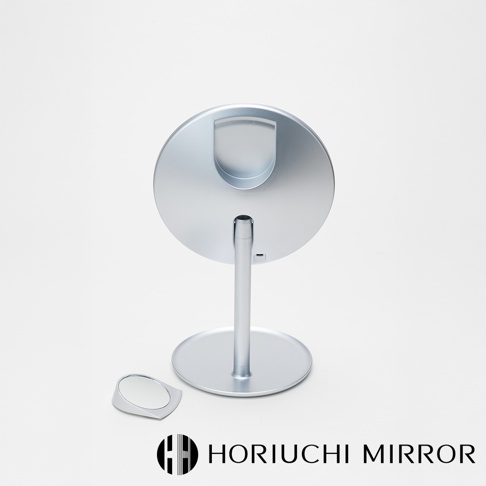 銀色金屬材質的桌上型化妝鏡，圓形鏡面，帶有支架和底座，鏡背有半圓形置物槽，旁邊放著一個小型三倍放大鏡。