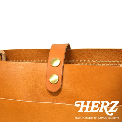 HERZ 品牌標誌和 HAND MADE IN JAPAN Since1973 字樣，搭配近拍的皮革包細節，包含兩個金屬鉚釘的皮帶扣。