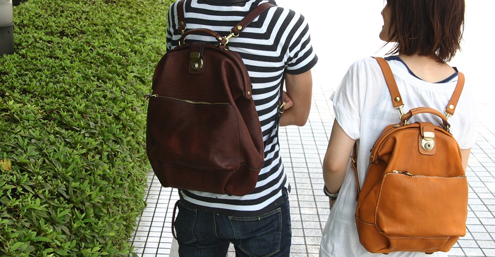 兩款職人手作皮革輕旅行後背包，一款為深棕色，另一款為焦糖棕色，皆為圓潤造型，配有頂部提把、前置拉鍊口袋及金屬鎖扣。