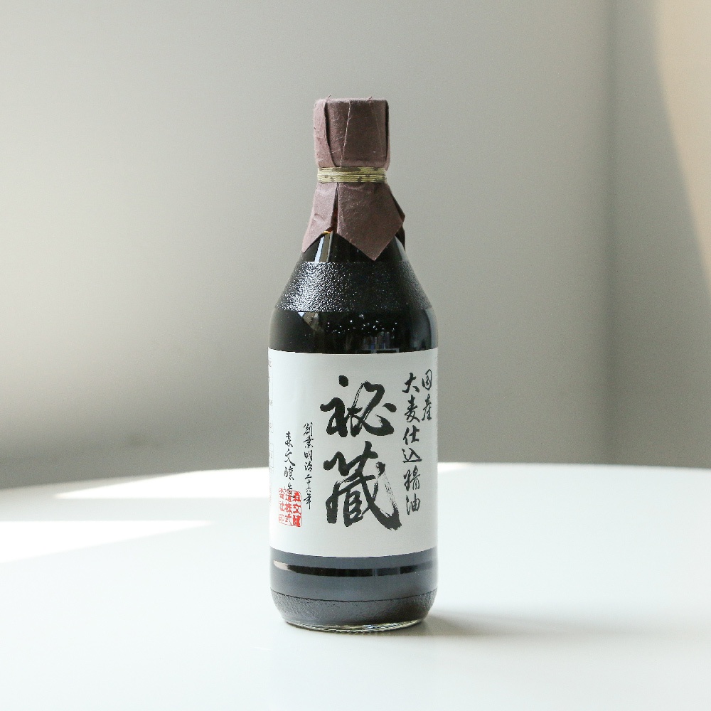 玻璃瓶裝著深色醬油，瓶頸處以紙質封套和金色細線裝飾，標籤為白色，印有黑色日文漢字。