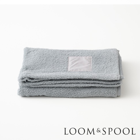 灰藍色純棉大浴巾疊放，上面有淺灰色標籤，下方有 LOOM & SPOOL 商標