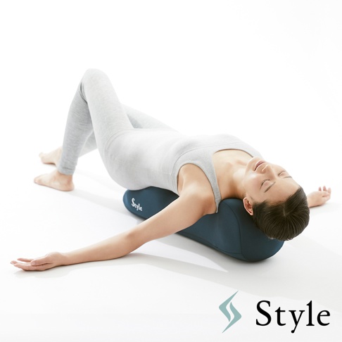 一名女子仰臥，頭部後仰，背部彎曲，躺在一個深藍色的長條形舒展棒上。舒展棒表面有「Style」字樣。女子身穿淺灰色瑜珈服。