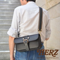 黑色職人手作皮革單扣經典斜背包，配有織帶和黃銅色金屬扣環，由皮革製成，斜挎於身穿條紋襯衫的男士肩上。