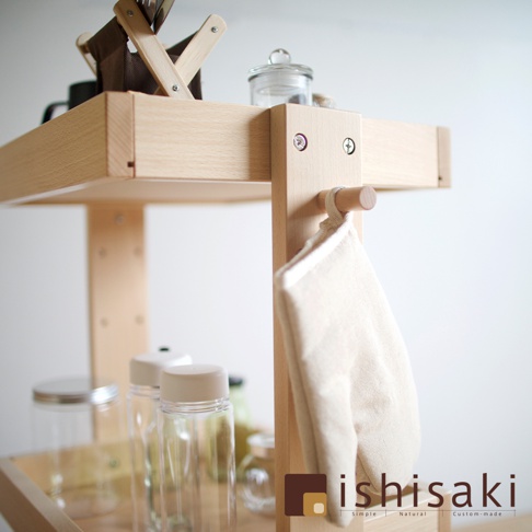 淺色木製收納推車，有兩層層架，側邊掛著米白色布料物品，上方層架擺放著玻璃罐和深棕色布料收納盒。