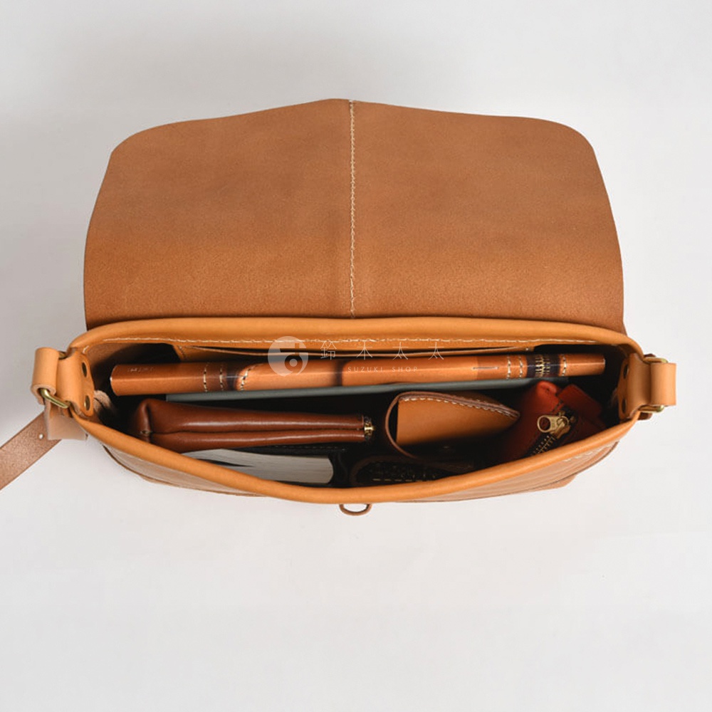 模式B： 從上方視角拍攝的復古棕色皮革馬鞍包，包蓋敞開，內部可見書籍、筆和一個紅色拉鍊皮夾。