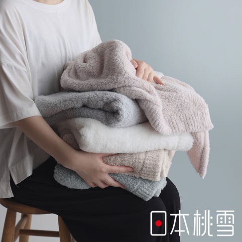 一位身穿白色上衣的人抱著一疊蓬鬆柔軟的毛巾，毛巾有粉紅色、灰色、白色和米色等多種顏色，背景為淺藍色牆面，右下角有「日本桃雪」的文字標記。