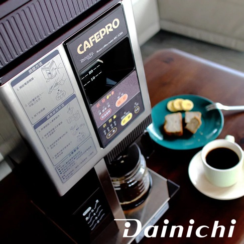 銀色和黑色 Dainichi CAFEPRO MC-520A 自動生豆烘焙咖啡機，設有黑色控制面板，顯示烘焙和研磨選項。旁邊有一個綠色盤子，上面有兩片吐司和香蕉片，還有一杯咖啡。