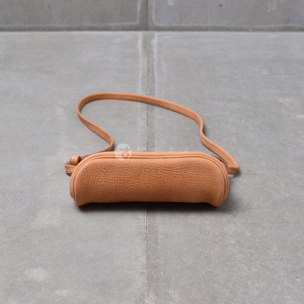 一個淺棕色皮革製的貼身斜背包，有著粗糙的紋理，斜背包平躺在灰色水泥地面上，一條細長的斜背帶從包包上方延伸開來。