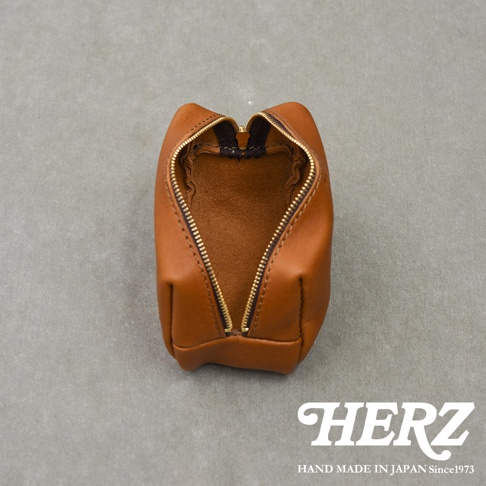一個呈現俯視視角的日本HERZ職人手作棕色皮革拉鍊收納包，內部為未經內襯處理的麂皮質感，配有精緻的金屬拉鍊，下方標註品牌名稱與日本手工製造資訊。