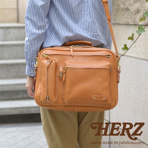 咖色皮革製成的職人手作兩用單肩包，設有拉鍊主隔層、多個外部拉鍊口袋及插袋，側面有可拆卸的拉鍊收納袋，搭配頂部提把和可調式肩帶。