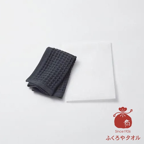 一件深灰色格紋毛巾，疊放在白色紙板旁，右下角有一個紅色印章圖案，上面寫著 Since 1926 和日文。