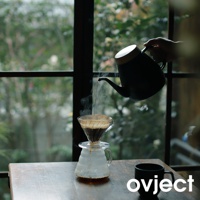 黑色琺瑯手沖壺，壺蓋為木質，正在向玻璃濾杯中沖煮咖啡，濾杯下方是玻璃分享壺，旁邊有一個黑色馬克杯，背景為窗外的綠意景致。