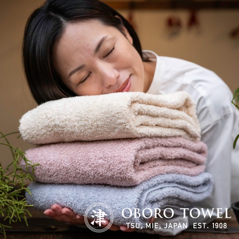 女性手持疊放的有機棉浴巾，顏色由上至下分別為米白色、淡粉色和淺藍色，浴巾質地柔軟蓬鬆。