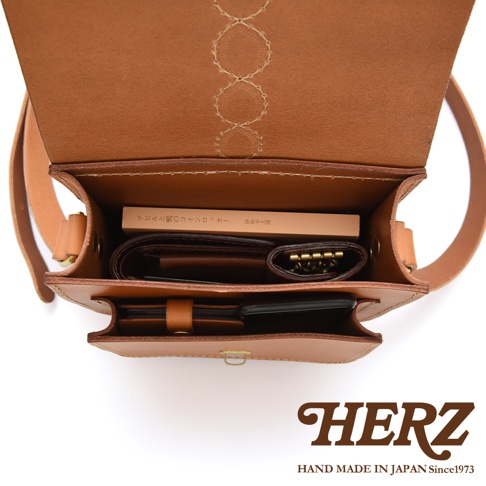從上方視角俯瞰日本HERZ手工皮革側背包的內部構造，展示其優異的收納空間。包內放置著短夾、鑰匙包與文庫本書籍，前置立體口袋則分類收納手機與隨身小物。皮革蓋內側精緻的十字繡裝飾與堅韌的原色牛皮質感清晰可見，整體設計簡約實用。