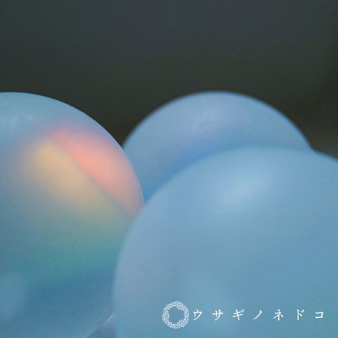 三個霧面藍色稜鏡球，其中一個球體內部有彩虹光暈，背景為深灰色，右下角有白色日文品牌標誌。