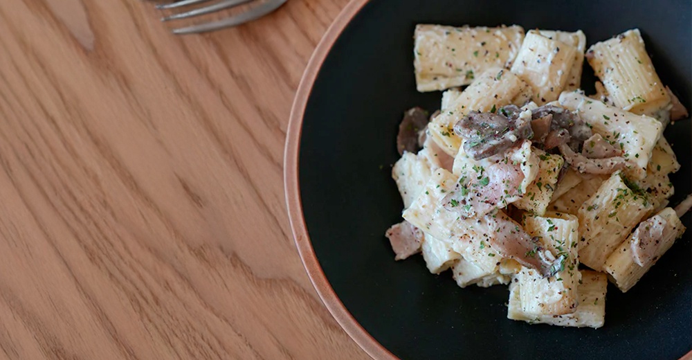 黑色陶瓷義大利麵盤盛著奶油培根義大利麵，撒有香料與黑胡椒，盤子邊緣帶有淺褐色陶土質感。