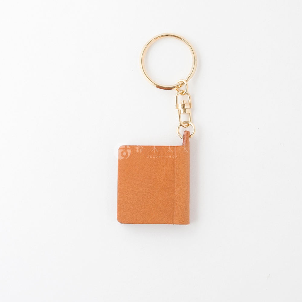 棕色皮革書本造型鑰匙圈，配有金色金屬環和掛鉤，皮革表面壓印有品牌標誌。