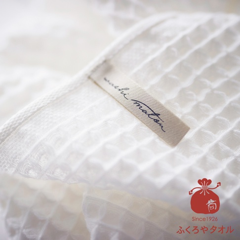 白色格紋織法的毛巾，上面縫著一條淺米色織標，織標上有藍色手寫字體「washi matou」。圖片右下方有一個紅色布袋圖案，下方有 Since1926 字樣及日文「ふくろやタオル」。