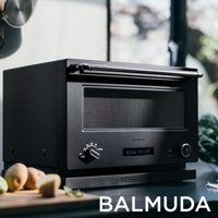 黑色BALMUDA The Range 20L微波烤箱，配有銀色把手，前方有黑色旋鈕、數位顯示螢幕及啟動按鈕。產品放置在黑色底座上，背景為模糊的室內景觀，右側有綠色蔬菜。