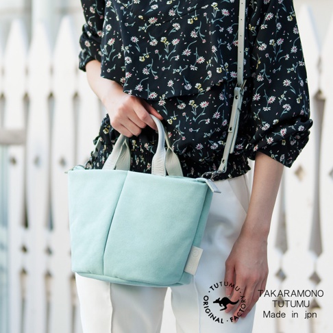 一位女士手提淺綠色麂皮水桶包，身上穿著印花襯衫，背景是白色木柵欄。