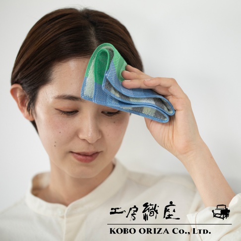 女性以手持藍綠色今治格紋信封式長方形手帕擦拭額頭，手帕材質為棉，格紋設計包含藍色、綠色與灰色。