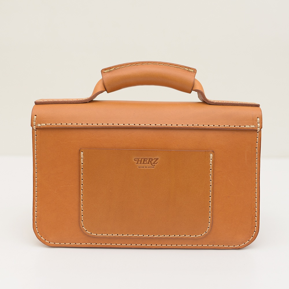 職人手作皮革雙扣兩用迷你肩背包，呈現溫潤的焦糖棕色調。包身由厚實的皮革製成，表面光滑，帶有細緻的手縫線條。正面有一個方形的貼袋，袋口處壓印著「HERZ」與「MADE IN JAPAN」的品牌標誌。包款設有圓潤的提把，結構穩固，整體散發著復古質樸的質感。