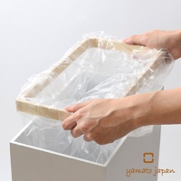 一雙手正將一個透明塑膠垃圾袋套入一個白色方形垃圾桶內，垃圾袋邊緣固定在一個木紋色擺動式掀蓋的框架上。